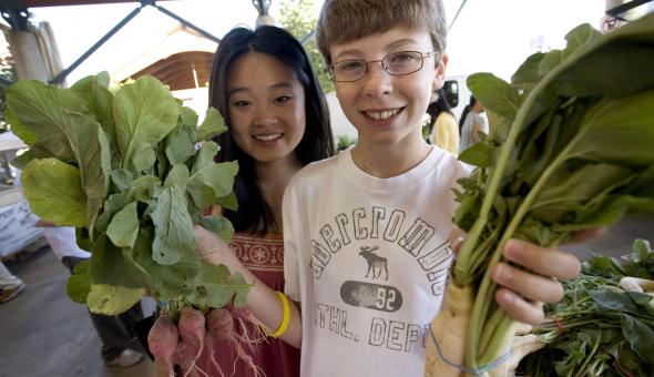 two kids at farmers marketing showing leafy greens to camera