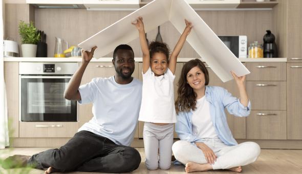 Family of three sitting on the kitchen floor, holding up an art board; child in the middle raises both arms to hold the v-shaped white board