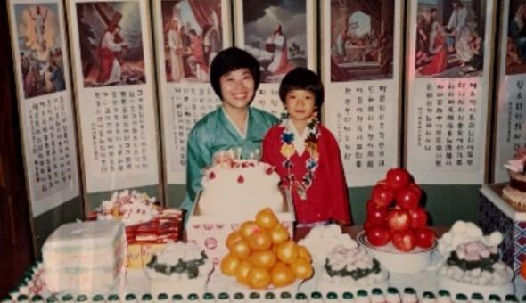 Peter Cho wearing red hanbok and flower garland with his mother, who is wearing teal hanbok at a table of colorful foods in front of a Lunar New Year display.