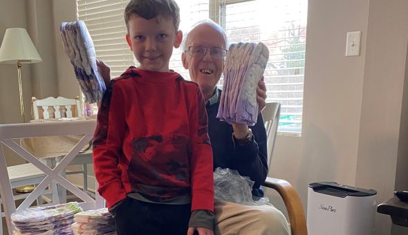 Jonas, an 8-year-old leans on the knee of his grandpa who is holding up bundles of diapers that they are wrapping for donation.