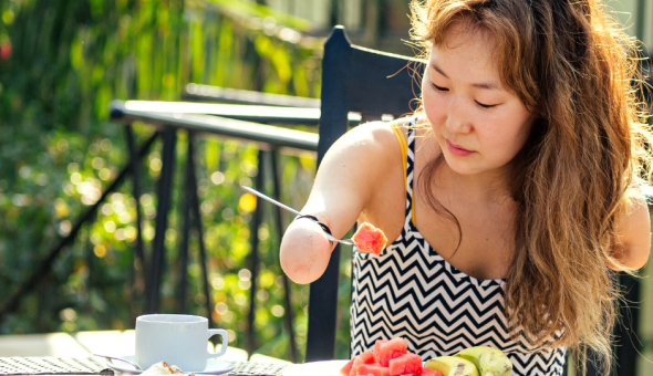 Teen with wavy hair eats watermelon outdoors with a loop fork on her upper arm.