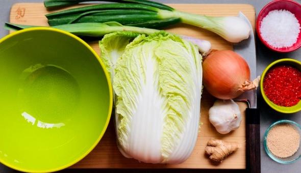A green bowl beside kimchi ingredients: napa, onion, garlic, scallions, salt, and ground chili