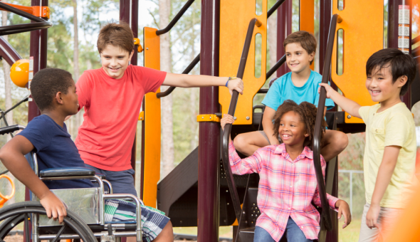 Five children are talking at a playground. Two are sitting on the playground, one is sitting in a wheelchair, and two are standing.