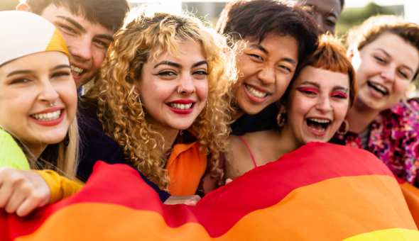 Seven young friends standing together outside celebrating LGBTQ+ Pride with a rainbow flag