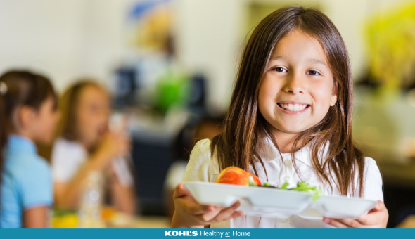 Student with brown hair holding a school meal tray with friends eating at a table behind her
