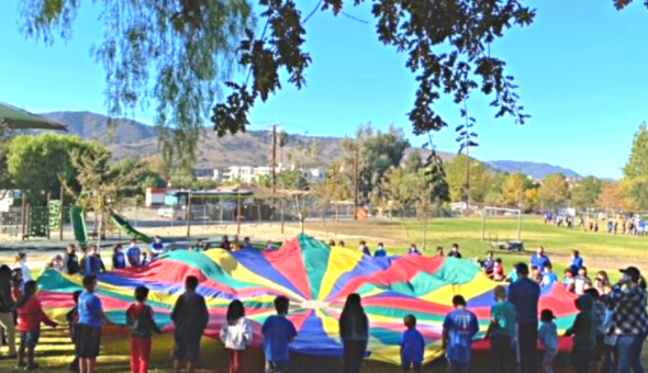 A group of Conejo Elementary students, families, and staff circle a giant rainbow parachute outdoors