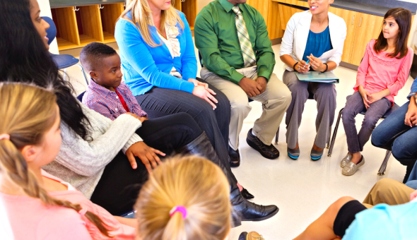 A group of school families sit on chairs in a circle getting to know each other. Four children and four adults are pictured, and two are partially in the picture.