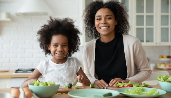 A woman and child smile together as they prepare a green salad and other vegetables on light blue plates.
