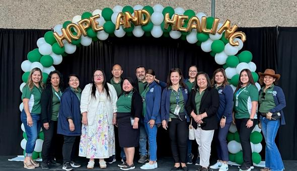 Parent University leadership team (14 adults) stands together on a stage under a green and white balloon arch with gold writing that reads "Hope and Healing"