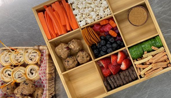 Two snack trays with snacks, including deli pinwheel wraps, carrot sticks, popcorn, berries, chocolate-covered pretzels, nut butter