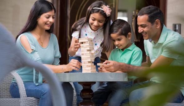 Family of two adults and two children play a block stacking game on patio.