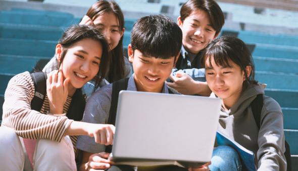 Five teens sit together in bleachers around a laptop on a sunny day, smiling.