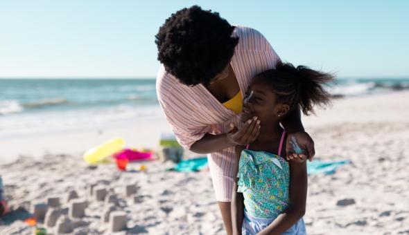 Mother and child apply sunscreen at the beach with the ocean in the background.