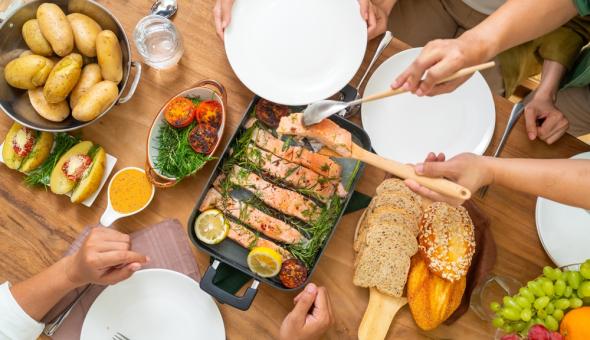 From above, a table with family serving each other portions of salmon, potatoes, bread, tomatoes, and side dishes.