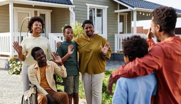 Two families wave hello outside a house. Six people of different ages are present and one person is using a wheelchair.